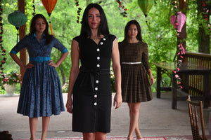 A woman wearing a fitted black pencil dress with white buttons, standing confidently in a modern setting. The dress is sleek, elegant, and flattering, blending business and style.